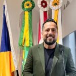 O vereador João Luiz Luz, presidente da Câmara Municipal de Biguaçu, em pose profissional, sorrindo e usando terno verde, diante de bandeiras oficiais do Brasil, Santa Catarina e do município.