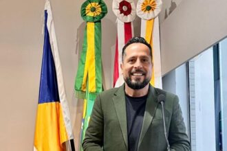 O vereador João Luiz Luz, presidente da Câmara Municipal de Biguaçu, em pose profissional, sorrindo e usando terno verde, diante de bandeiras oficiais do Brasil, Santa Catarina e do município.