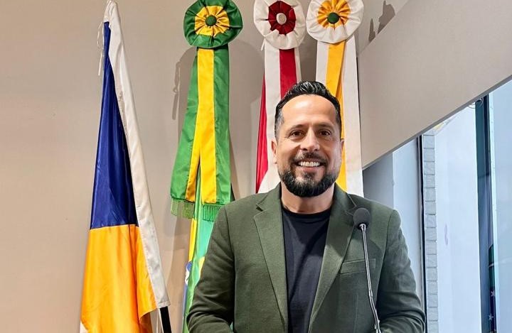 O vereador João Luiz Luz, presidente da Câmara Municipal de Biguaçu, em pose profissional, sorrindo e usando terno verde, diante de bandeiras oficiais do Brasil, Santa Catarina e do município.